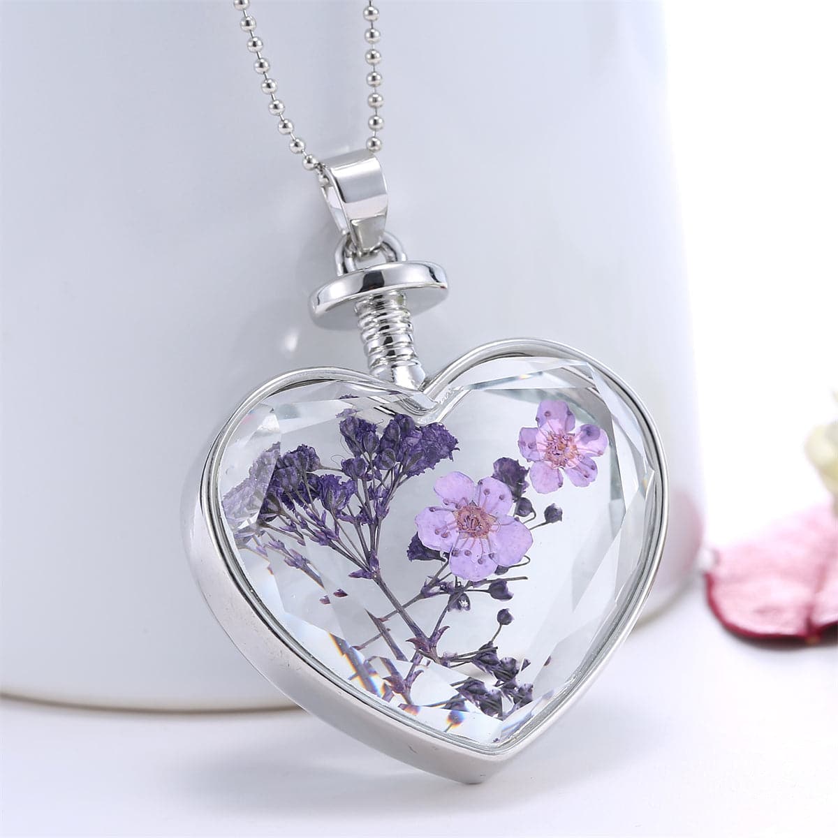 Heart-shaped silver pendant necklace featuring pressed purple peach blossoms encased in resin. A delicate floral accessory.