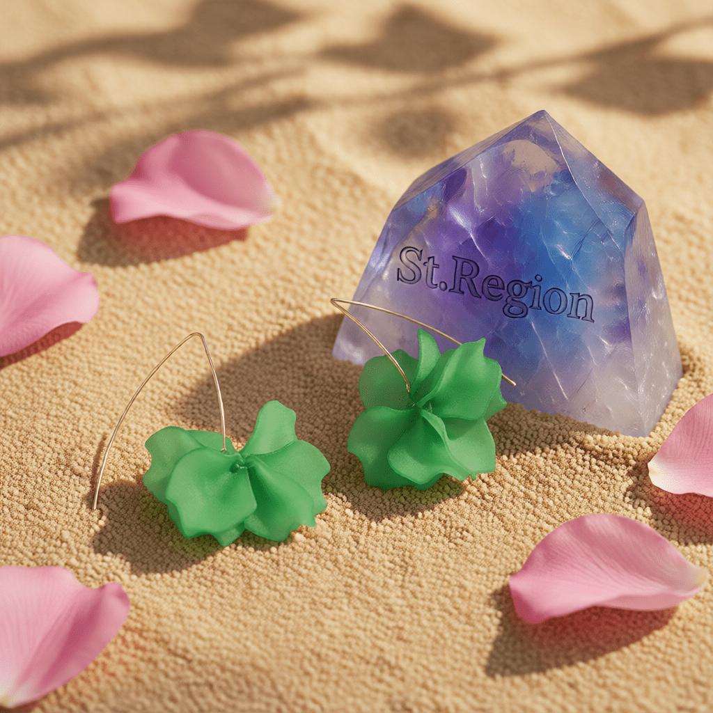 Green flower-shaped earrings with thin wire hooks, displayed on a sandy surface with pink flower petals and a translucent purple crystal labeled 'St. Region'.