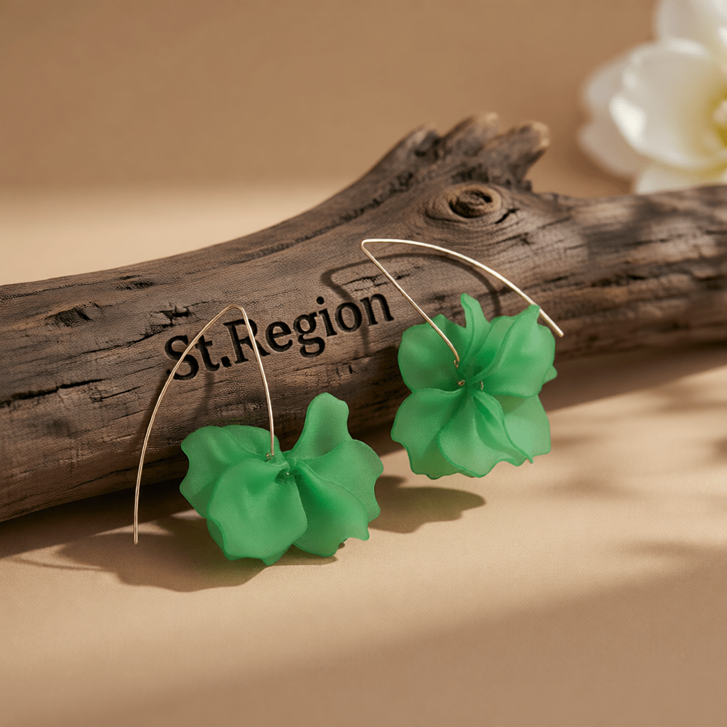 A pair of green flower-shaped drop earrings with thin gold hooks, displayed on a weathered wood branch over a beige surface.