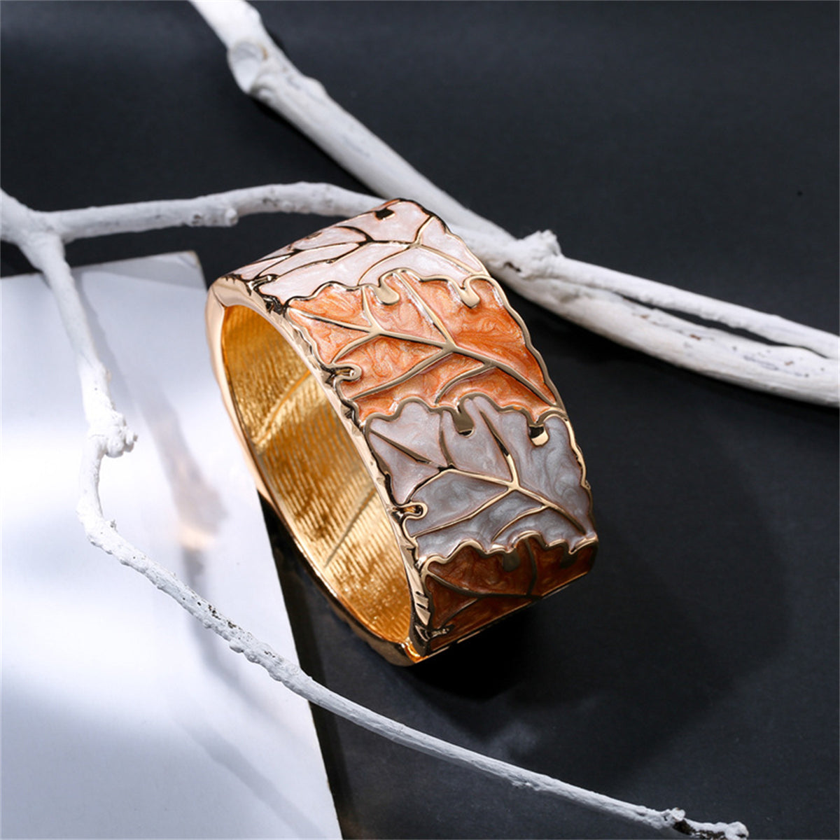 A stylish bangle in gold and white, designed with a leaf pattern, complemented by orange enamel and 18K gold plating.